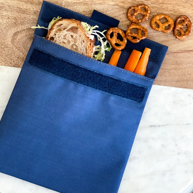 Reusable sandwich and snack bag shown with a sandwich, carrots and pretzels. velcro closure and insulated interior. Durable material. Navy Blue. wipe clean or throw in washing machine.