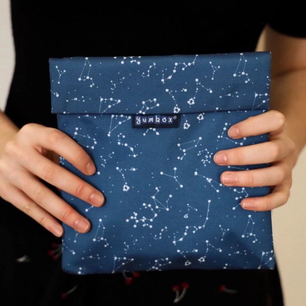 Woman holding a reusable sandwich and snack bag.  velcro closure and insulated interior.  Durable material.  white constellations on blue background.  wipe clean or throw in washing machine.