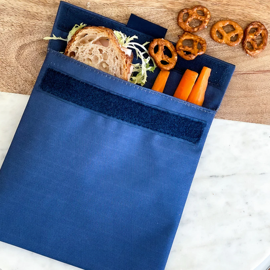 Reusable sandwich and snack bag shown with a sandwich, carrots and pretzels.  velcro closure and insulated interior.  Durable material.  Navy Blue.  wipe clean or throw in washing machine.