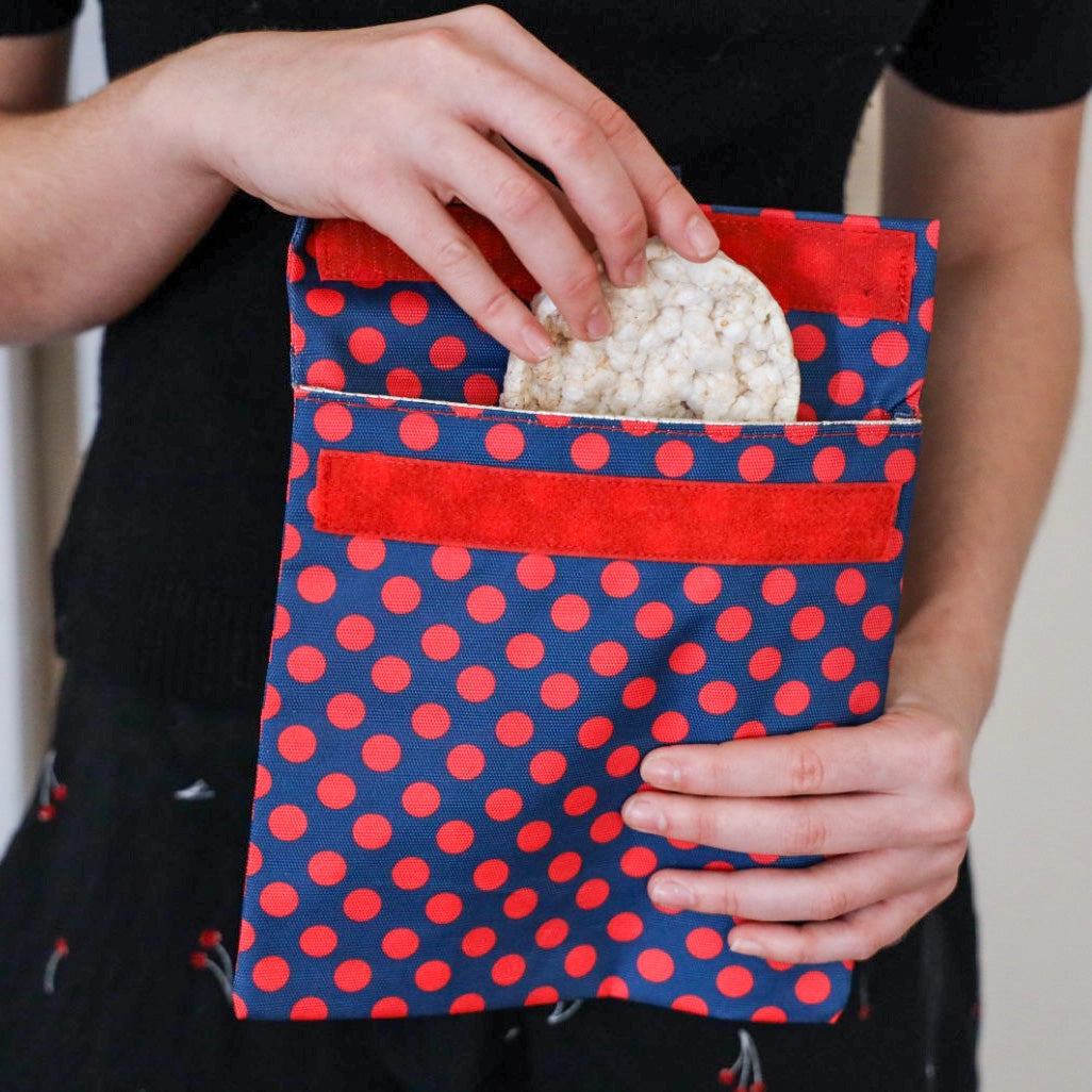 Woman holding  a reusable sandwich and snack bag.  velcro closure and insulated interior.  Durable material.  red polka dots on dark blue background.  wipe clean or throw in washing machine.
