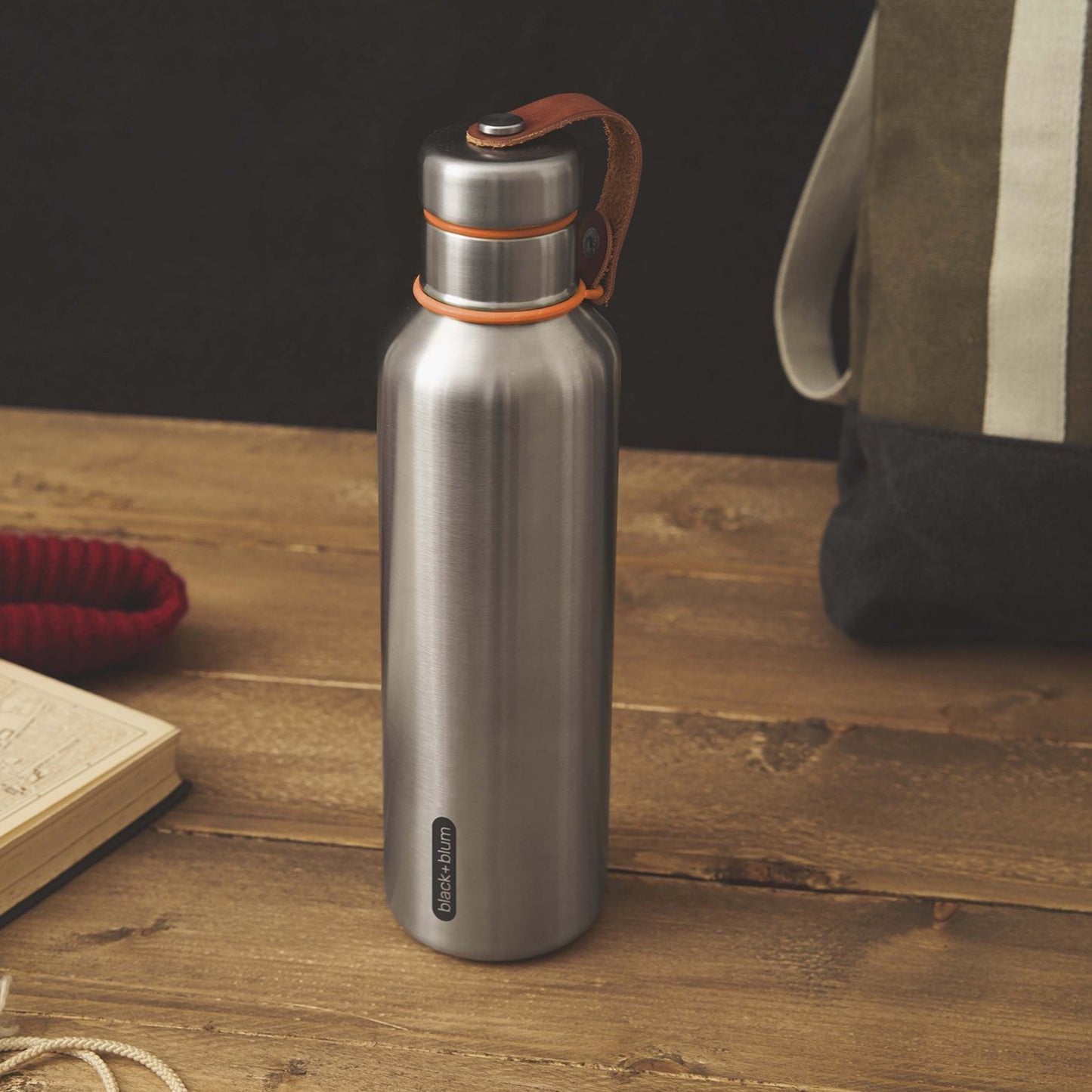 Insulated stainless steel water bottle with vegan leather accent and orange trim.  On wood table by bag and book